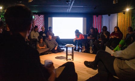 A group of Somali people sit in a circle around a cassette player on a table in the centre of a darkened room 