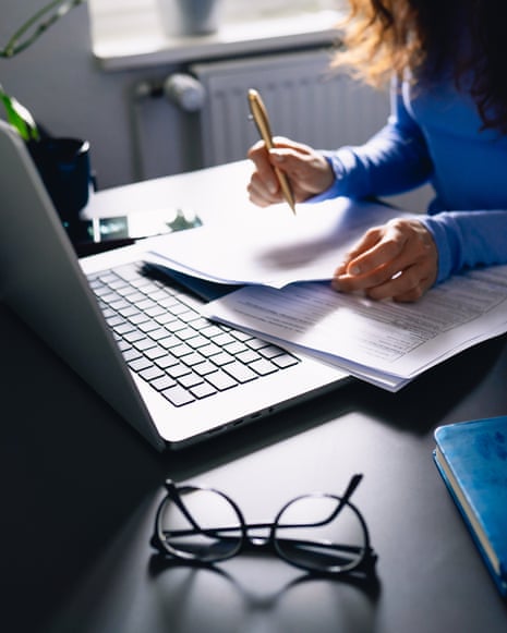 A woman's hands write on tax paperwork beside an open laptop on a desk