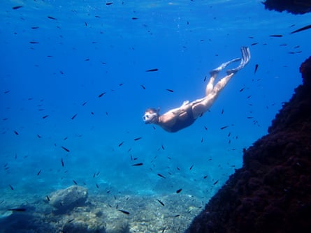 A woman wearing flippers, diving mask and swimming costume swimming in bright blue water surrounded by small fish