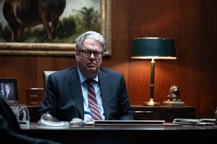 Wearing a suit, he sits behind a large, dark desk in a moodily lit office