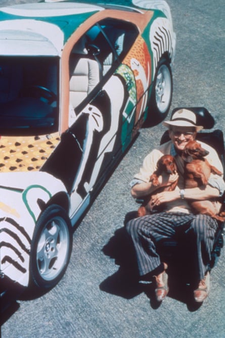 Artist David Hockney sits in a chair with his sausage dogs beside a painted car