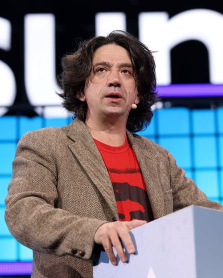 Wearing a brown jacket over a red T-shirt, with a microphone attached to his head, he stands at a lectern on stage