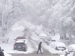 A person crosses a road lined with snow-covered cars and trees