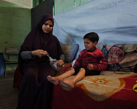 A woman wearing a black headscarf applies saline solution to her young son who sits beside her. He is crying.