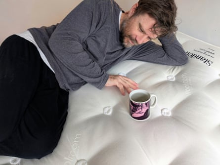 Man with a beard in a long-sleeved shirt over a T-shirt lies on a bare mattress with his head on his hand and a mug of water in front of him