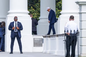 Donald Trump in a blue suit and white baseball cap walks between white pillars. A security agent in a suit and sunglasses stands looking away from Trump