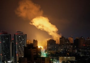 A street lined with high-rise buildings with a cloud of smoke rising above them, and in the distance a bright yellow flash of light at ground level