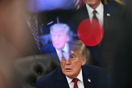 Donald Trump sits at the resolute desk as a digital image of him seen through an out-of-focus broadcast camera hovers slightly above his head