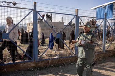 A soldier with a rifle standing by a high metal fence topped with barbed wire. On the other side of the fence are dozens of women and a few men