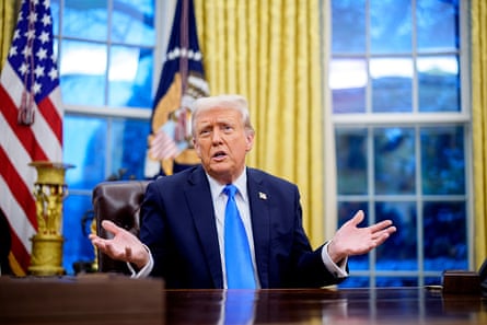 Donald Trump mid-speech sitting at his large brown desk at the White House, wearing a dark-blue suite, white shirt and blue tie, with his arms bent and his hands out to the side