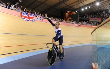 Sir Chris Hoy waves while riding a bike in a velodrome