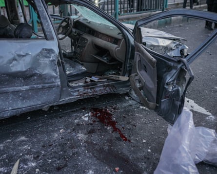 The open driver-side door of a destroyed car reveals blood that has run down from the driver's seat