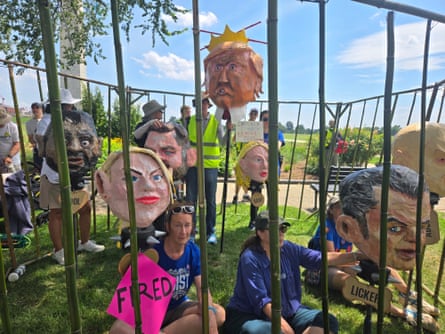 group stands and sits in fake cage on grass while some hold up oversized heads of trump and other administration figures