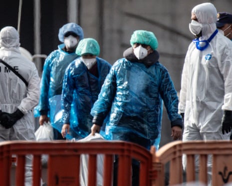 People wearing blue plastic ponchos, hair coverings and face masks carry large plastic bags after disembarking the ship