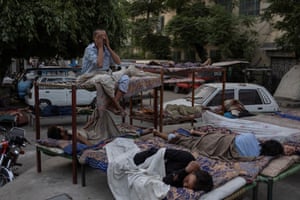 People rest on makeshift beds in an outdoor setting amid parked cars