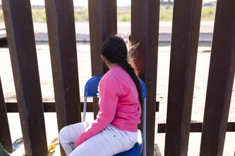 A child sitting on a chair looks through a metal fence.