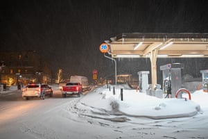 Cars pass by the gas station at night. Snow covers the road while the sidewalk is buried in deep snow