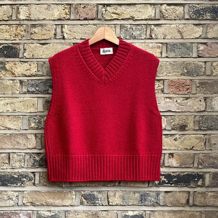 British wool V-neck vest from the brand Elwin at Gather & See. The vest is red and hangs on a wooden hanger against a light stone brick wall.
