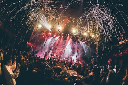 In a dark club lit by stage lights, streamers explode over a busy, boisterous crowd while a band dance and sing on stage