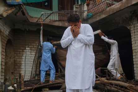 A man covers his face with his hands and weeps in a damaged house. Two other men can be seen moving debris behind him