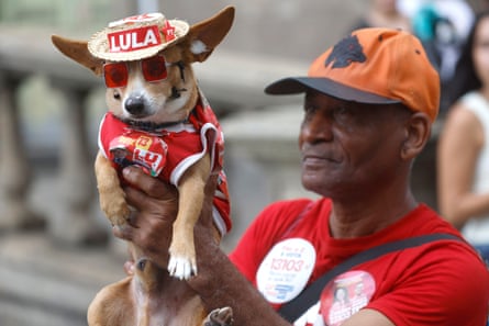 Man in red T-shirt holding up a dog with cowboy hat saying Lula on it and wearing a Lula T-shirt.