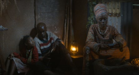 A man and a girl sit on the floor of a dark hut lit by a paraffin lamp and talk as a solemn woman in a headdress sits before a large mortar