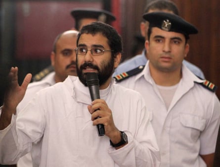 Alaa Abd el-Fattah speaks into a microphone; he has a uniformed guard behind him. He has a dark beard and wears a white shirt and small spectacles.