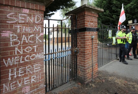 A message reading ‘Send Them Back Not Welcome Here Never!’ has been chalked on a brick gatepost at the site. Police officers stand guard at the gate and someone is holding an England flag behind them