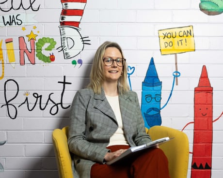 Melissa Anderson a woman in a blazer jacket, top and red long skirt, sitting in a yellow chair against a white wall with an artistic mural painted on it