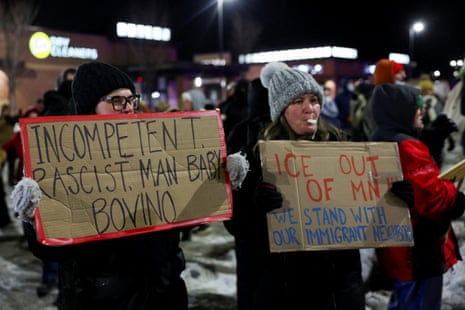 Two women, dressed in full winter gear, hold up signs. One sign reads "incompetent racist man baby Bovino" while the other reads "ICE out of MN we stand with our immigrant neighbors"