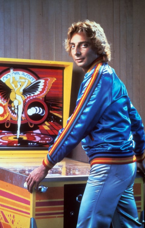 Barry Manilow wearing a shiny blue jacket with orange and yellow trim looks over his shoulder while playing a pinball machine