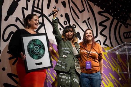 Emily Wurramara wearing a linen suit with slogans including “climate change” and “care for country”, with one fist holding a microphone raised, and her arm around Shellie Morris.