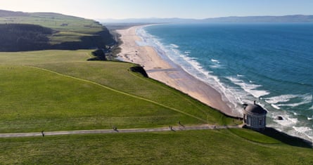 A round temple at the edge of a flat green clifftop with a beach and the sea below