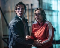 Sam Claflin in navy Barbour-type jacket and Kaley Cuoco in red and white track suit top looking sweaty and frightened at something off camera - clutching at each other's arms