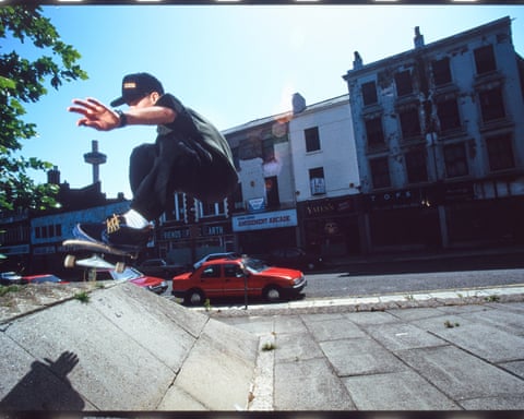 A skateboarder mid-trick above a paved area, with a red car and weathered buildings in the background