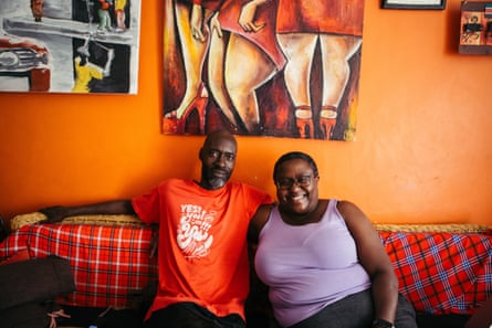 A smiling middle-aged African man and woman sit together on a sofa with paintings behind them on an orange wall