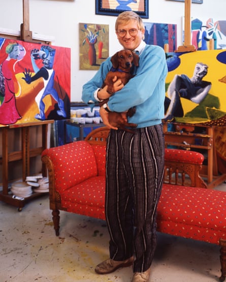 Hockney stands holding one of his dogs, with colourful paintings behind, including 2 on easels