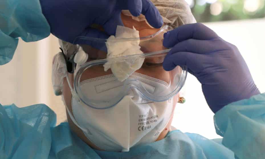 A healthcare worker wearing a sterile gown, gloves, mask, and hair covering wipes the inside of their goggles with a cloth.