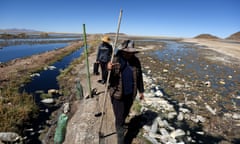 Two Indigenous Andean women walk along a path on a raised ground beside a rubbish-strewn lake