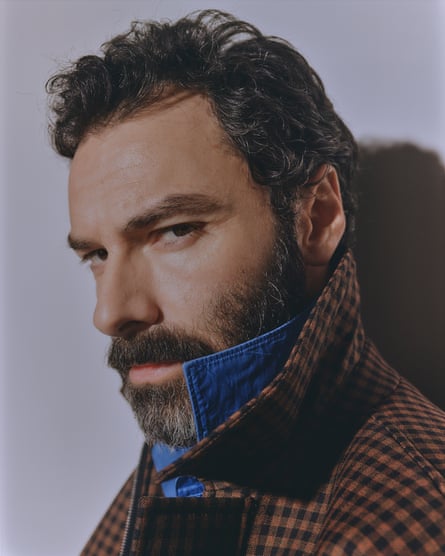 Aidan Turner looks sideways, wearing a checked jacket and a blue shirt, both with turned-up collars