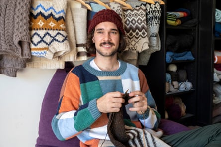 Australian knitter Brendan Girak, knitting, with jumpers hanging behind him. March 2021
