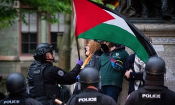 Helmeted and armored police confront a young woman waving a Palestinian flag.