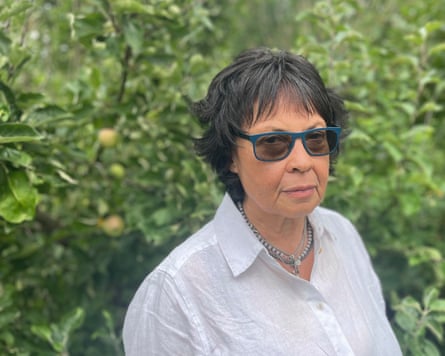 Joyce Thomas stands outdoors in front of greenery. She has short dark hair cut in a fringe and wears a white linen shirt and blue-rimmed sunglasses.