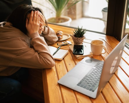 A young woman using a laptop slouches on the table and covers her face with her hands.