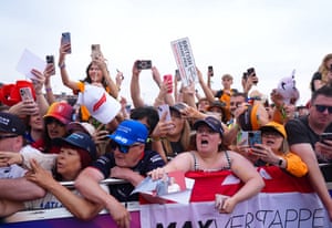 A crowd of people pressing against barriers hold up phone cameras and look towards someone or something out of frame. Some of them are wearing hats in the colours of different F1 teams