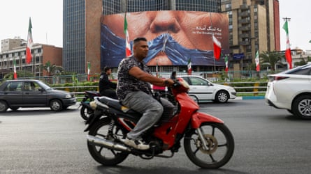 Man on motorbike driving past the billboard, which shows the strait of Hormuz like a strip of scrunched-up fabric, sewn across Trump’s mouth