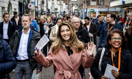 Dilan Yeşilgöz-Zegerius wearing a pink coat smiles at the camera while walking among crowds in a street