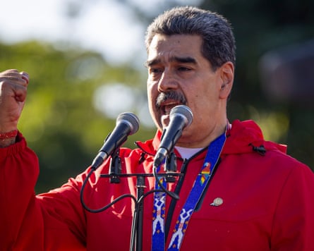 Nicolás Maduro wearing a red jacket speaking into a microphone and raising his hand.