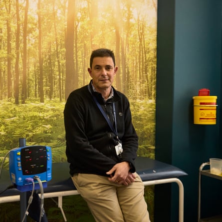 A man wearing a lanyard sits next to hospital equipment against a wall decorated with a forest mural