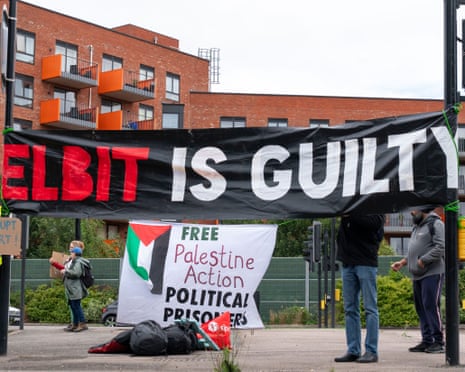 A large black banner reading ‘Elbit is guilty’ held between lampposts hangs above a smaller ‘Free Palestine Action political prisoners’ banner